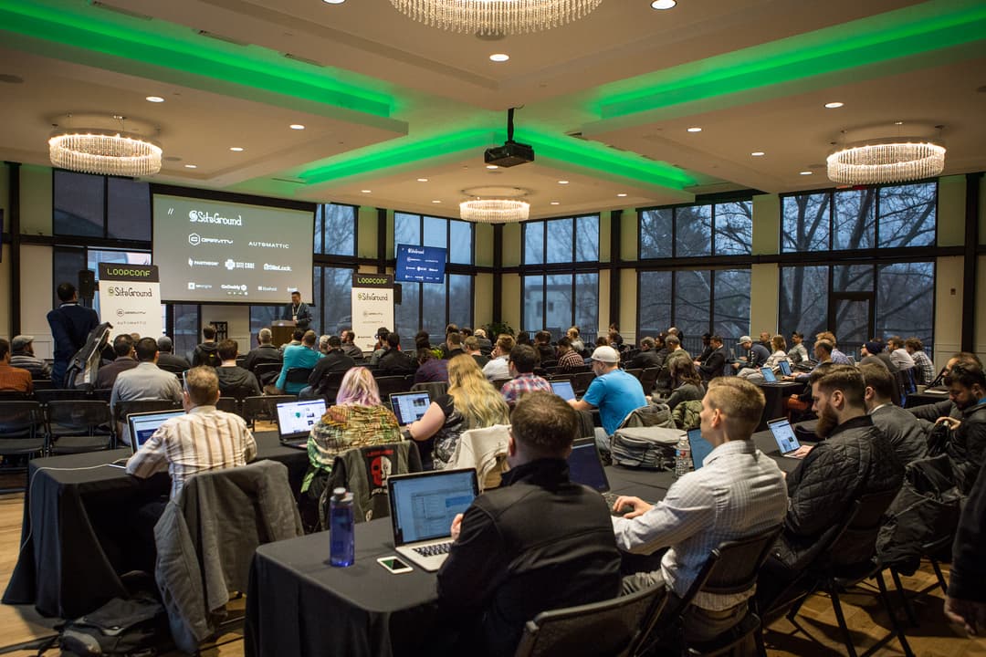 A crowd of people at a LoopConf event, some sitting and others standing, with a speaker on stage in the distance.